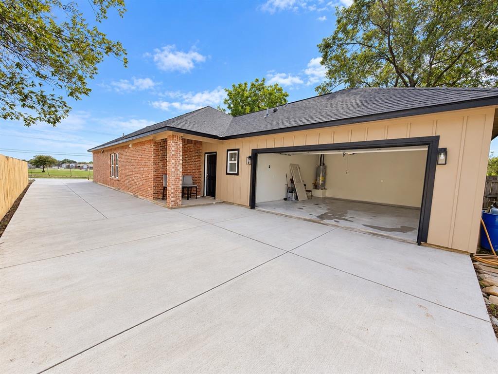 Exterior featuring a brick and board-and-batten siding facade, two-car garage with an open bay, extended concrete driveway, covered porch with a brick pillar, and shingle roofing