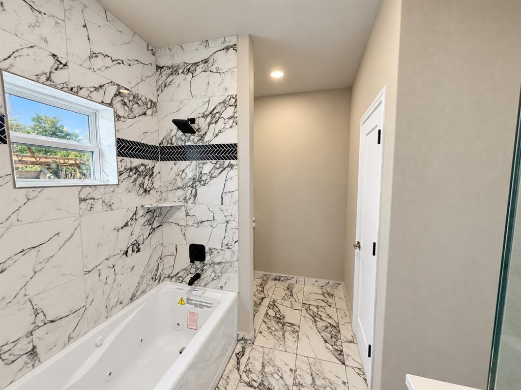 Jetted soaking tub with large-format veined tile surround, matte black rainfall shower fixture, decorative herringbone tile inlay, and matching stone-patterned tile flooring