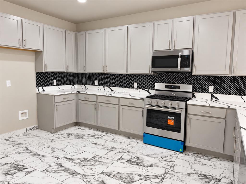 Kitchen featuring light gray shaker-style cabinetry, patterned stone-finish countertops, black herringbone tile backsplash, stainless steel appliances, and large-format veined tile flooring Kitchen featuring light gray shaker-style cabinetry, patterned stone-finish countertops, black herringbone tile backsplash, stainless steel appliances, and large-format veined tile flooring