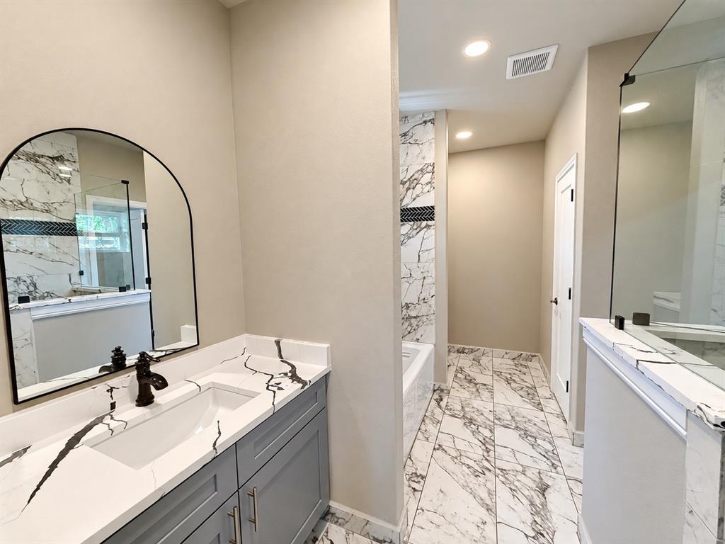 Bathroom featuring gray shaker-style cabinetry, white countertops with dark veining, an arched wall mirror, large-format tile flooring, and a walk-in shower with glass enclosure