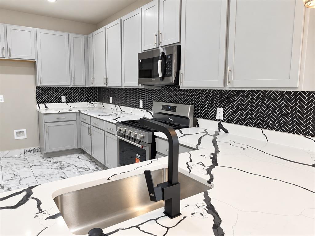 Kitchen featuring gray shaker-style cabinetry, high-contrast herringbone tile backsplash, white stone-patterned countertops, stainless steel appliances, and a matte black gooseneck faucet