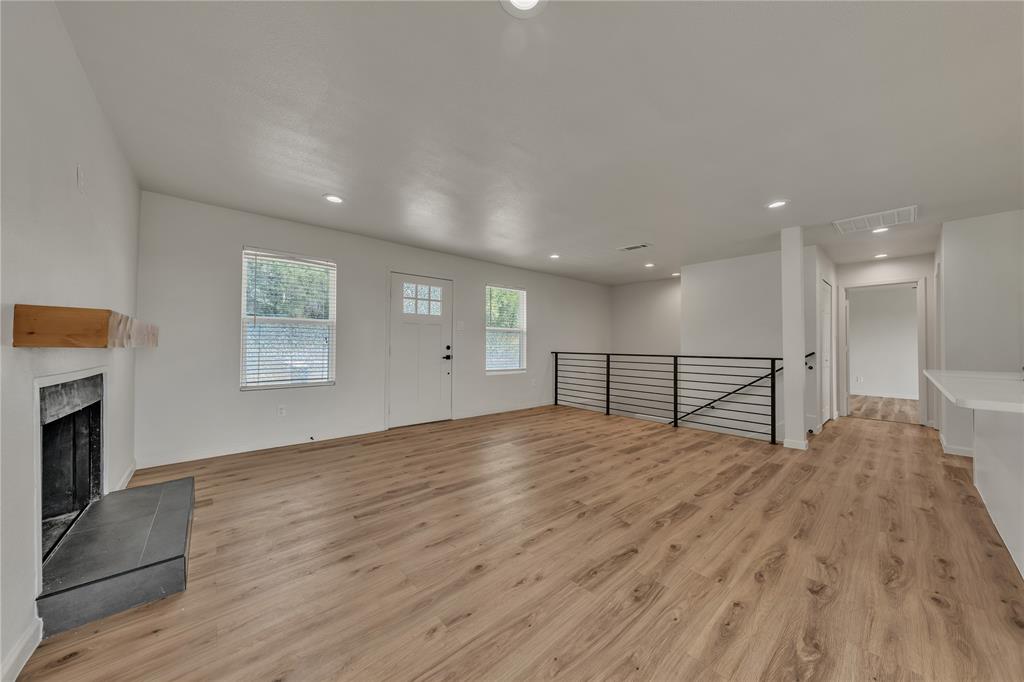 Open-concept interior featuring wood-finish flooring, recessed lighting, a stone-surround fireplace with a timber mantel, and an industrial-style metal stair railing Open-concept interior featuring wood-finish flooring, recessed lighting, a stone-surround fireplace with a timber mantel, and an industrial-style metal stair railing