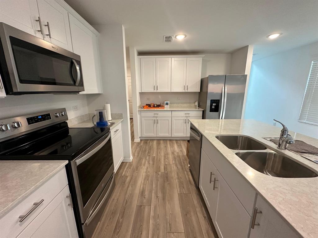 Kitchen featuring white Shaker-style cabinetry, stainless steel appliances, a central island with a double-basin sink, quartz countertops, and wood-finish flooring
