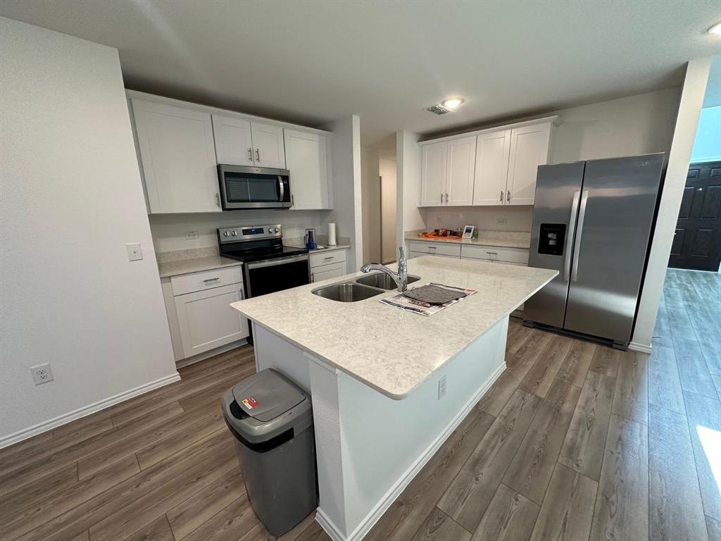 Kitchen featuring white shaker-style cabinetry, stone-finish countertops, stainless steel appliances, a center island with an integrated double-basin sink, and wood-finish flooring Kitchen featuring white shaker-style cabinetry, stone-finish countertops, stainless steel appliances, a center island with an integrated double-basin sink, and wood-finish flooring