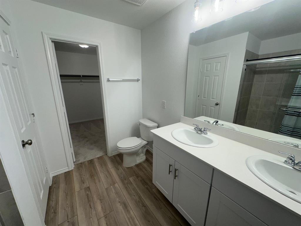Bathroom featuring a dual-sink vanity with a solid-surface countertop, white cabinetry with brushed metal hardware, wood-finish flooring, and a full-width mirror Bathroom featuring a dual-sink vanity with a solid-surface countertop, white cabinetry with brushed metal hardware, wood-finish flooring, and a full-width mirror