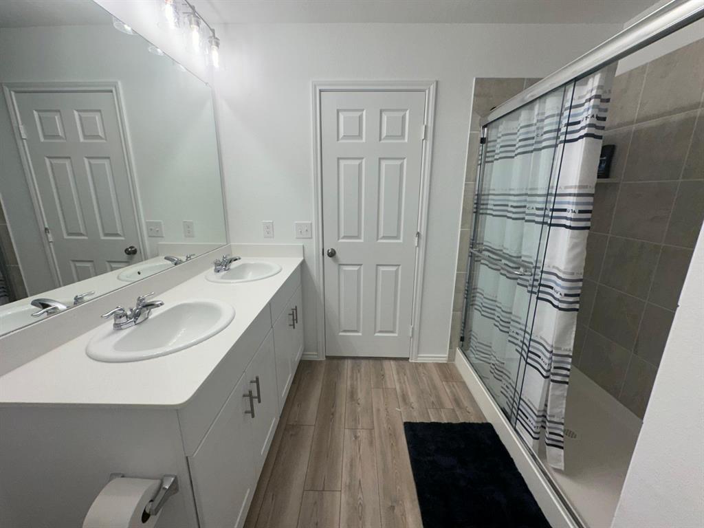 Dual-sink vanity with white countertop, full-width vanity mirror, wood-finish flooring, and glass-enclosed shower featuring tiled surround