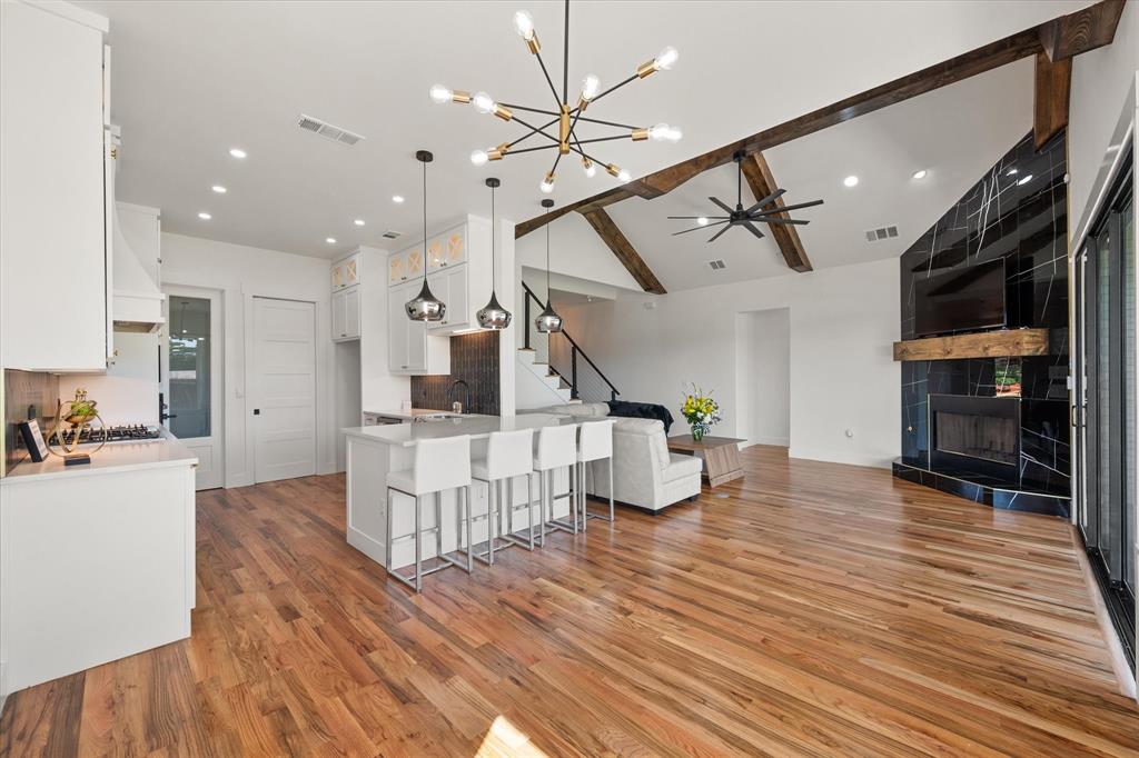 Open-concept interior featuring vaulted ceilings with decorative wood beams,Oak wood flooring, a kitchen island with pendant lighting, and a floor-to-ceiling fireplace with a black tile surround and a wood mantel