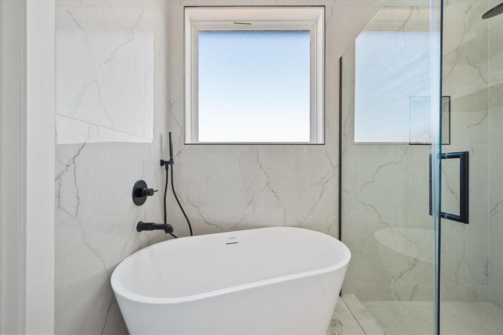 Freestanding soaking tub featuring matte black plumbing fixtures, oversized large-format tile surround, glass-enclosed shower, and built-in wall niche