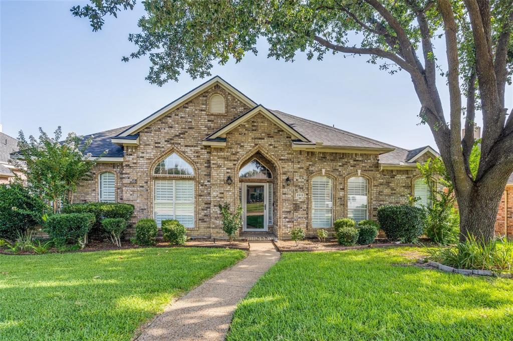 Brick exterior featuring arched window detailing, a steep-pitched roofline, and a concrete pathway leading to the front entrance, set amidst a landscaped front yard with mature trees