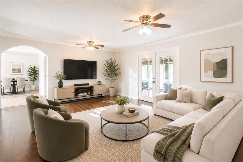 Living area featuring wood-finish flooring, two ceiling fans with light kits, crown molding, and double glass-paned doors leading to exterior space, highlighted by an arched doorway Living area featuring wood-finish flooring, two ceiling fans with light kits, crown molding, and double glass-paned doors leading to exterior space, highlighted by an arched doorway