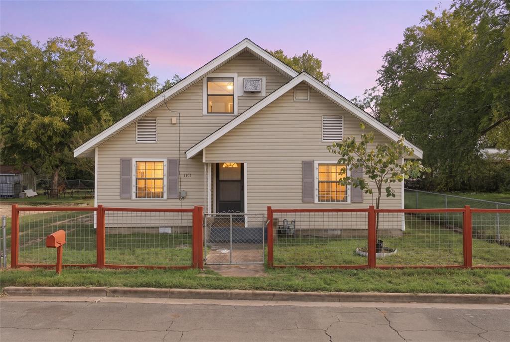 Exterior featuring light-toned horizontal siding, a gabled roofline, symmetrical shuttered windows, and a fenced front yard with a metal gate