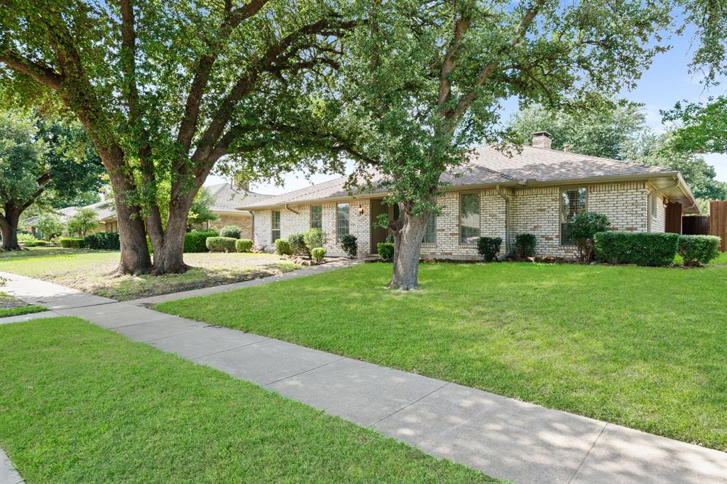 Single-story brick exterior featuring a hipped roofline, mature trees, manicured lawn, and a concrete sidewalk