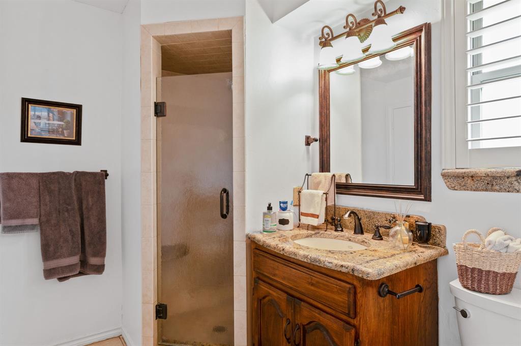 Bathroom featuring a wood vanity with stone countertop, drop-in sink, framed mirror with integrated lighting, and a glass-enclosed shower