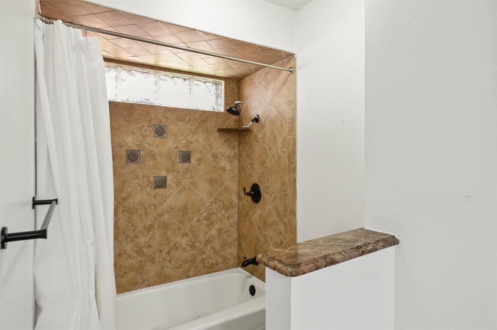 Shower and bathtub combination featuring diagonal stone-finish wall tile, decorative metal accent inlays, a built-in corner shelf, glass block window, and dark metal hardware
