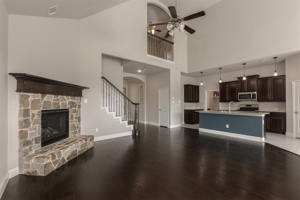 High-ceiling interior featuring a floor-to-ceiling stone fireplace with a wood mantel, dark wood-finish flooring, a tiered kitchen island with quartz countertops, custom wood cabinetry, and an open loft landing with metal balusters