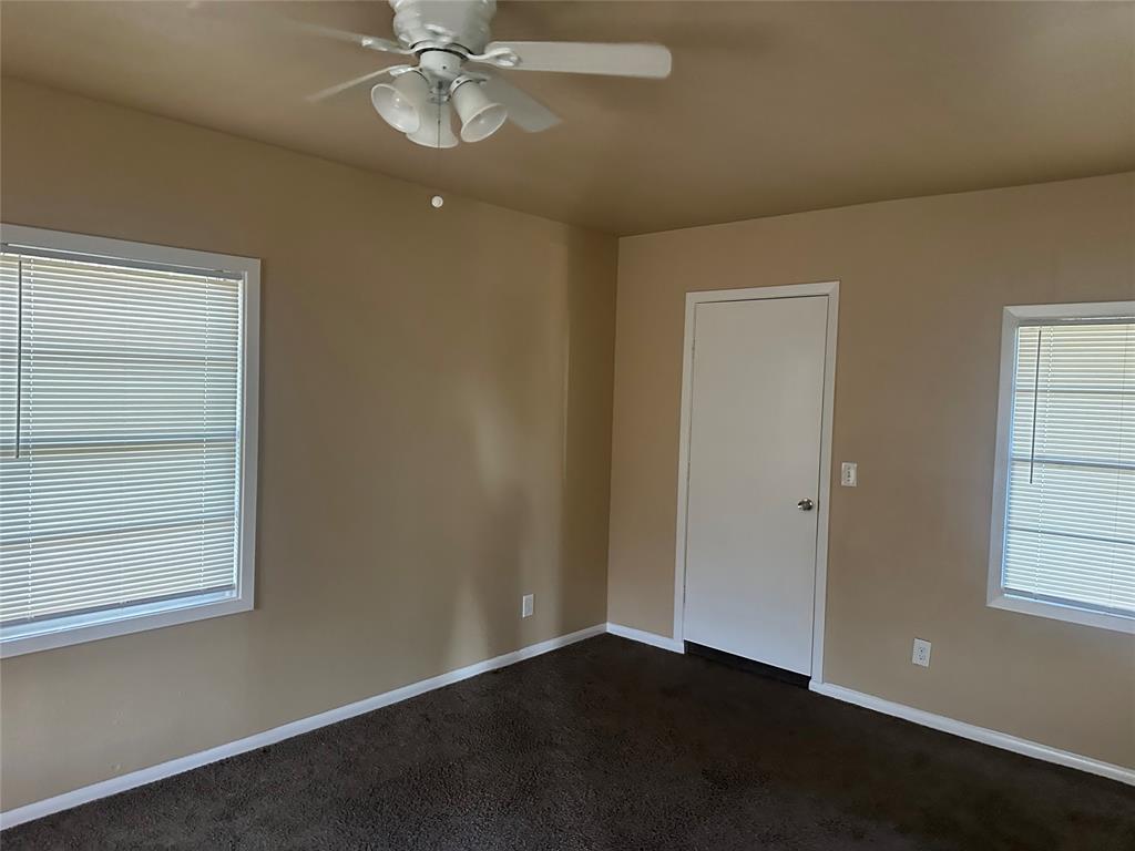 Neutral-toned room featuring dark carpeting, two windows with horizontal blinds, and a white ceiling fan with integrated lighting Neutral-toned room featuring dark carpeting, two windows with horizontal blinds, and a white ceiling fan with integrated lighting