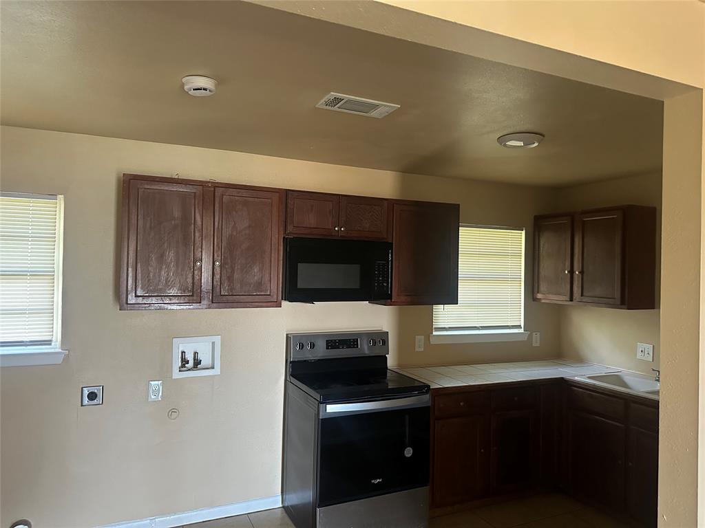 Kitchen featuring dark wood cabinetry, integrated black microwave, stainless steel range, tiled countertops, and a drop-in sink Kitchen featuring dark wood cabinetry, integrated black microwave, stainless steel range, tiled countertops, and a drop-in sink