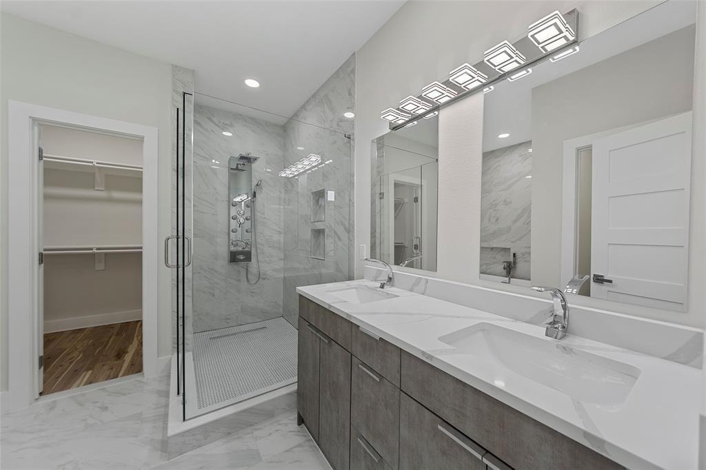 Dual-sink vanity with white stone-surface countertop, walk-in shower with multi-head fixture, stone-style tile flooring, and integrated wall-to-wall mirrors