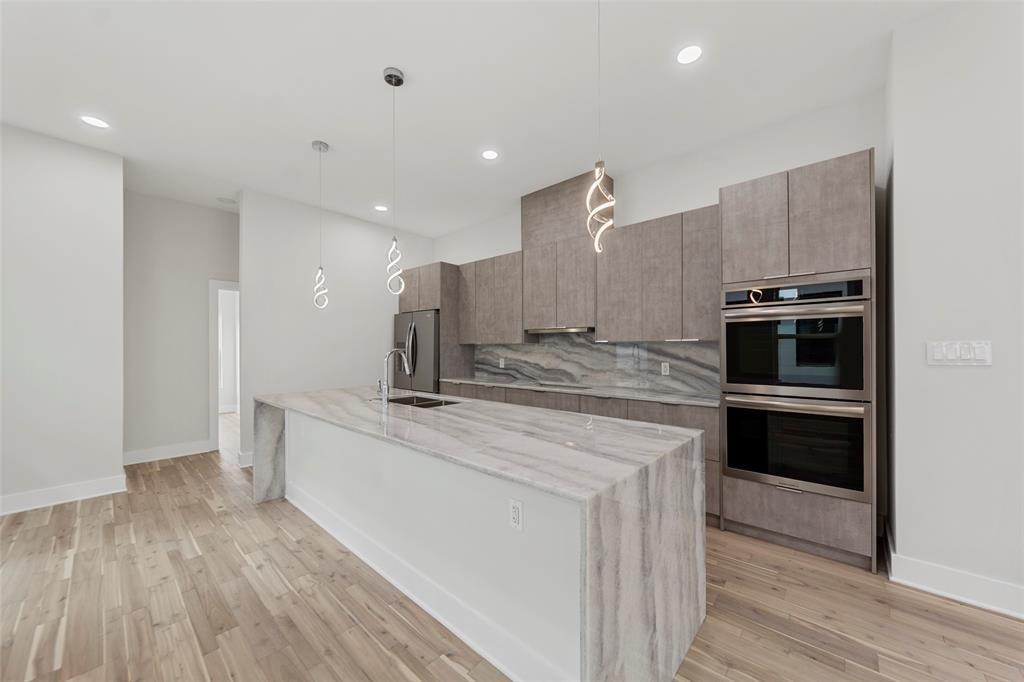 Kitchen featuring a waterfall-edge island with stone surfacing, stainless steel double wall ovens, wood-finish cabinetry, patterned stone backsplash, and pendant lighting