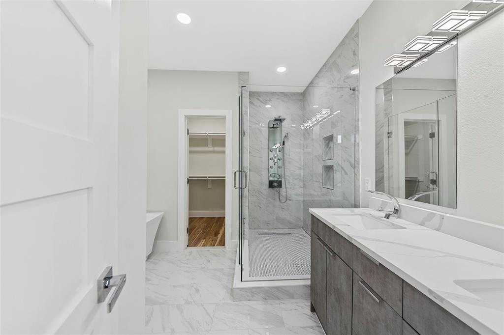 Bathroom featuring a dual-sink vanity with a solid-surface countertop, a walk-in shower with floor-to-ceiling stone-pattern tile, a ceiling-mounted shower panel system, and recessed lighting