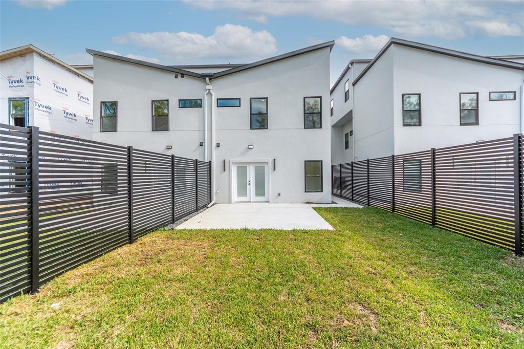 Modern two-story exterior featuring light-toned stucco siding, a sharp shed roofline, black horizontal slatted perimeter fencing, a concrete rear patio, and dual-pane glass entry doors