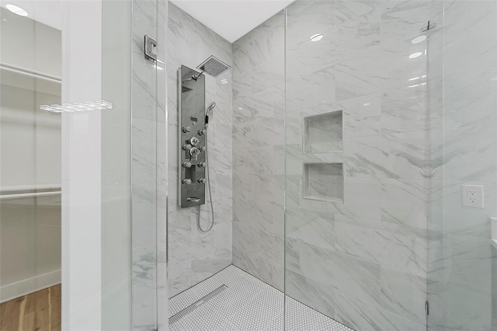 Walk-in shower featuring floor-to-ceiling stone-finish tile, recessed wall shelving, a stainless steel multi-head rainfall shower panel, and a mosaic tile floor with linear drain, enclosed by a frameless glass partition