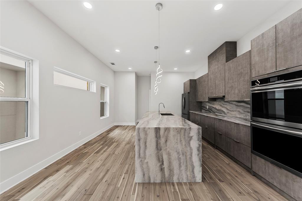 Kitchen featuring an expansive waterfall-edge island, textured cabinetry, stone-patterned backsplash, wood-finish flooring, and integrated stainless steel double wall ovens