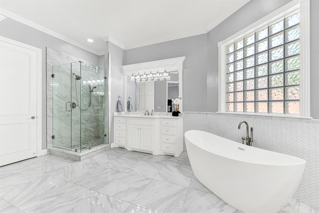 A seamless frameless glass shower features refined stone tile surrounds that complement the herringbone-patterned wall accents. Picture starting your mornings with a warm, refreshing shower before stepping out onto cool marble-look floors.