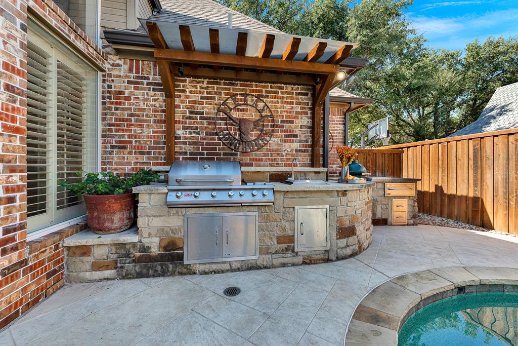 A built-in stone grill station sits sheltered under a custom timber arbor along the brick exterior wall. Picture sizzling up fresh steaks and appetizers while chatting with friends gathered poolside on warm summer evenings.