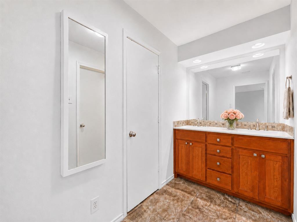 Double vanity featuring stained wood cabinetry, a solid surface countertop, an integrated tiled backsplash, and a large-format mirror with recessed lighting