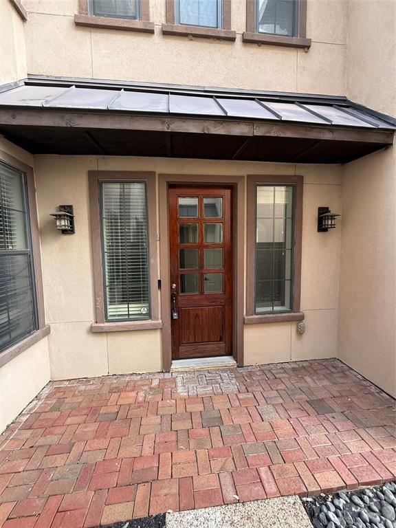 Stucco exterior entry featuring a brick-paved patio, a metal-roofed awning, a wooden eight-lite front door, and wall-mounted exterior sconces Stucco exterior entry featuring a brick-paved patio, a metal-roofed awning, a wooden eight-lite front door, and wall-mounted exterior sconces