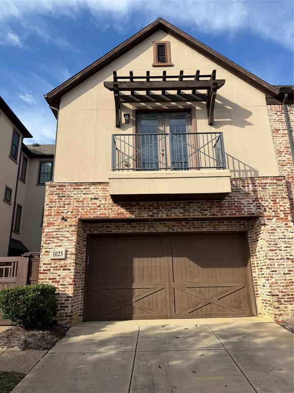 Stucco and brick facade featuring a second-story balcony with a metal railing, wood-beam pergola, and attached two-car garage with X-brace detailing