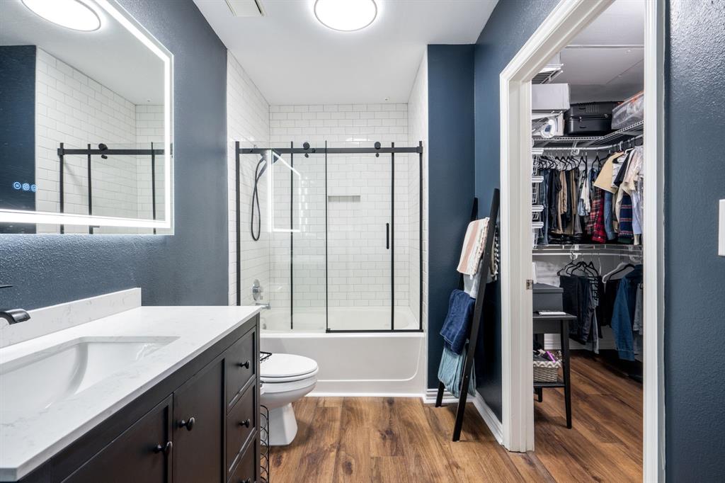 Bathroom featuring a vanity with a white countertop, an illuminated mirror, wood-finish flooring, a tub-shower combination with a black-framed sliding glass enclosure, and white subway tile wall surrounds