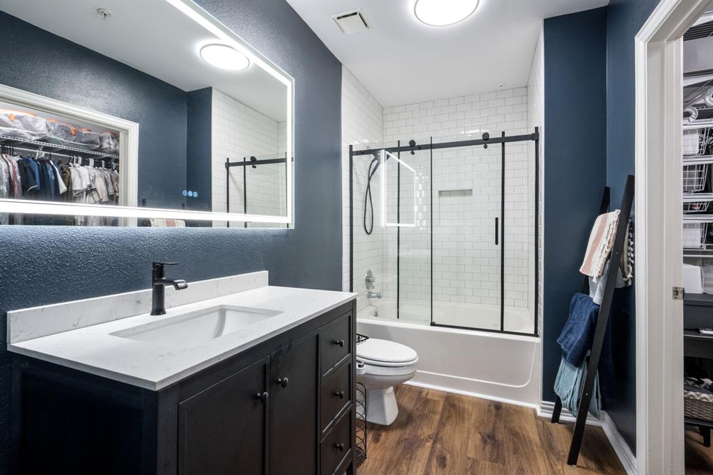 Bathroom featuring a dark wood vanity with a white stone-composite countertop, an integrated LED-backlit mirror, wood-finish flooring, and a bathtub with white subway tile surround and a sliding glass door with a black frame