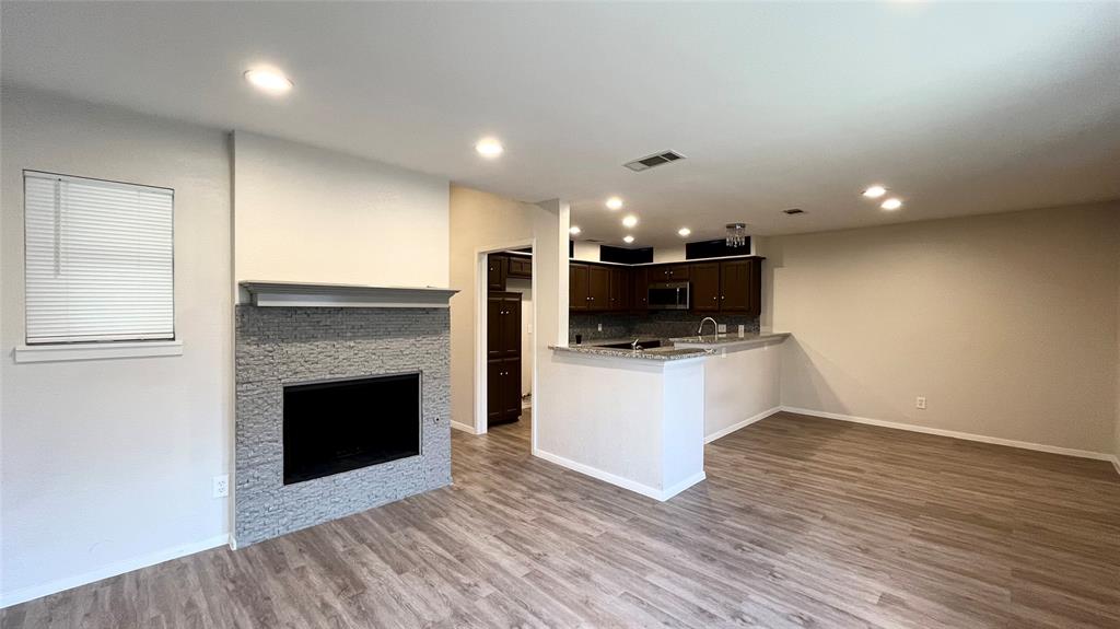 Fireplace with stacked-stone surround and mantel, wood-finish flooring, recessed lighting, and kitchen featuring dark cabinetry and a peninsula Fireplace with stacked-stone surround and mantel, wood-finish flooring, recessed lighting, and kitchen featuring dark cabinetry and a peninsula