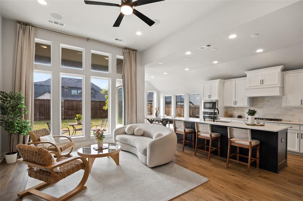 Open-concept living space featuring high ceilings with recessed lighting, oversized windows, wood-finish flooring, and a kitchen with white cabinetry and a contrasting center island Open-concept living space featuring high ceilings with recessed lighting, oversized windows, wood-finish flooring, and a kitchen with white cabinetry and a contrasting center island