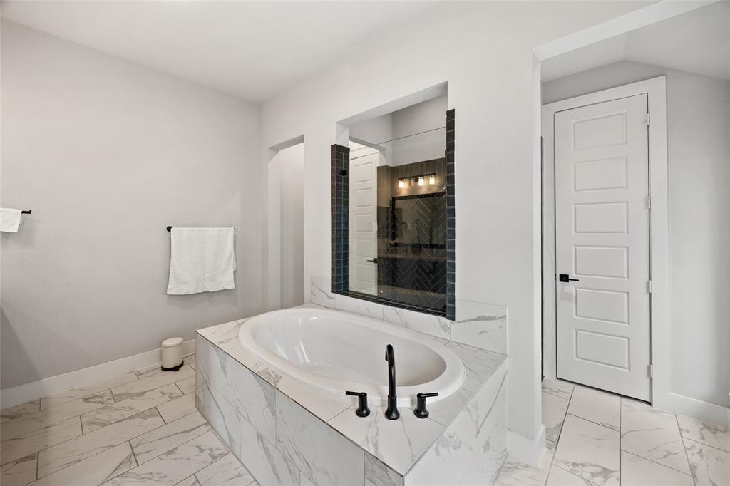 Oval soaking tub set in a stone-finish deck with a matte black faucet, surrounded by large-format stone-finish floor tile, a tiled accent wall with integrated mirror, and a five-panel solid core door