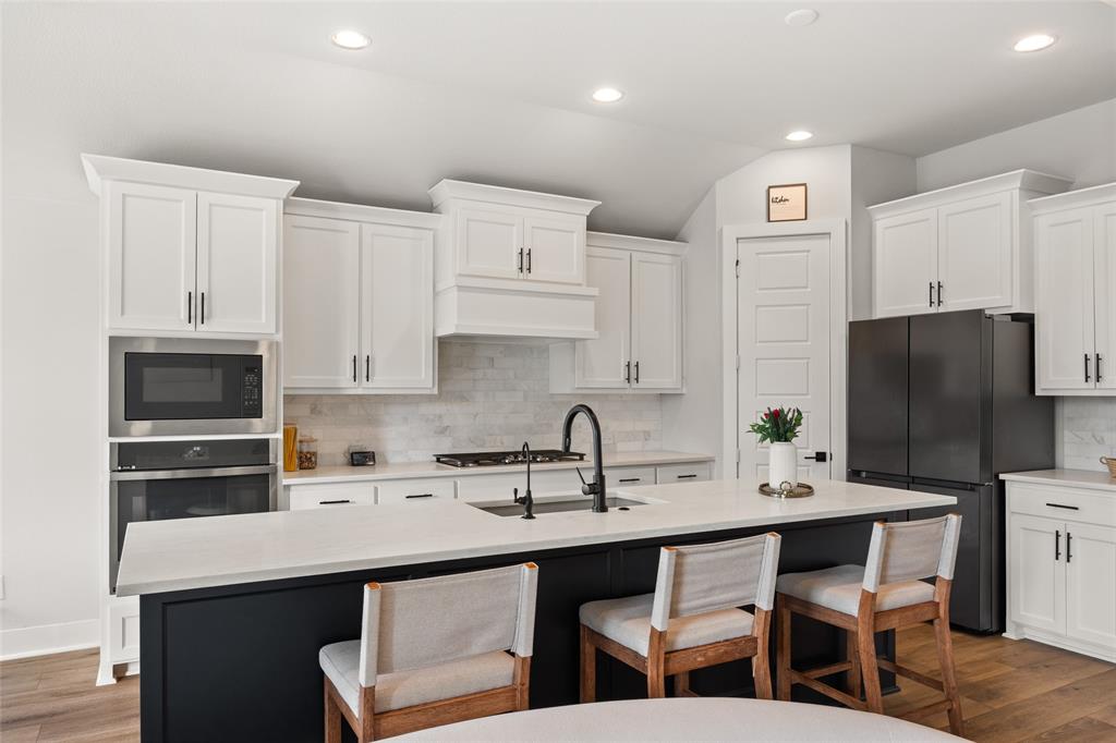 Kitchen featuring white shaker-style cabinetry, a contrasting dark island with a quartz countertop, stone tile backsplash, built-in stainless steel appliances, and wood-finish flooring