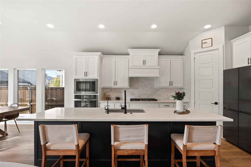 Kitchen featuring white shaker cabinetry, a contrasting dark kitchen island with a stone countertop, built-in wall oven and microwave, stone tile backsplash, and recessed lighting Kitchen featuring white shaker cabinetry, a contrasting dark kitchen island with a stone countertop, built-in wall oven and microwave, stone tile backsplash, and recessed lighting