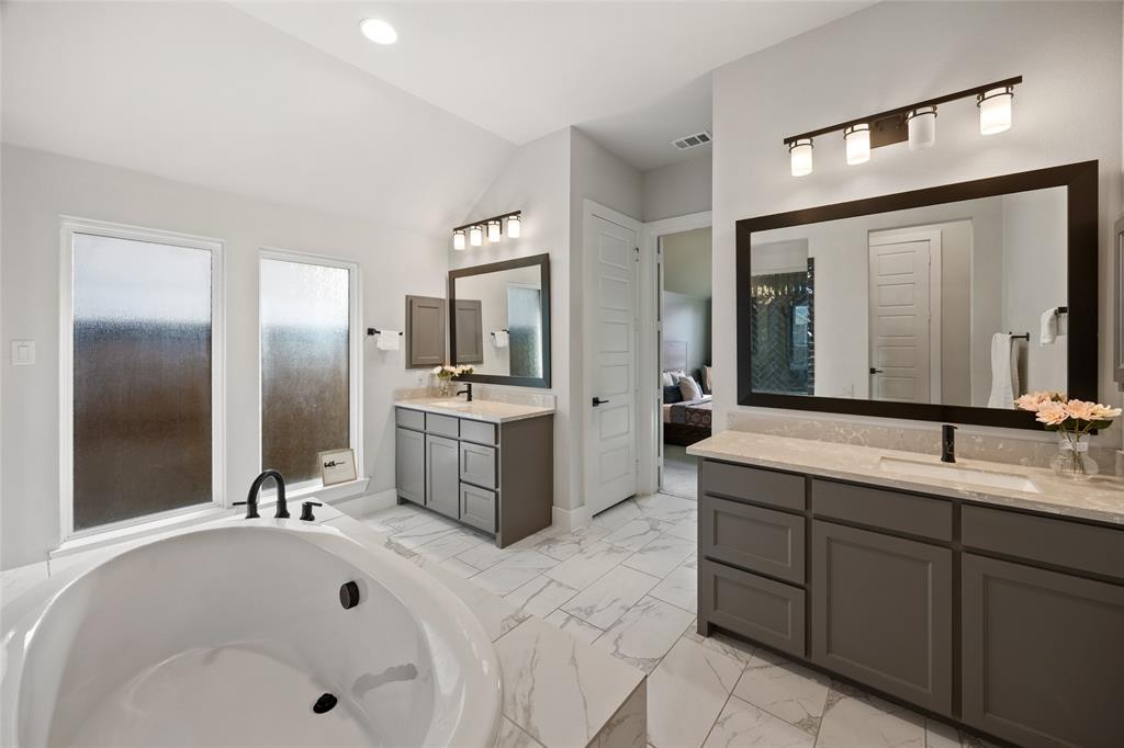 Dual vanities with shaker-style cabinetry, stone-finish countertops, tile flooring, freestanding soaking tub, and contemporary wall-mounted light fixtures