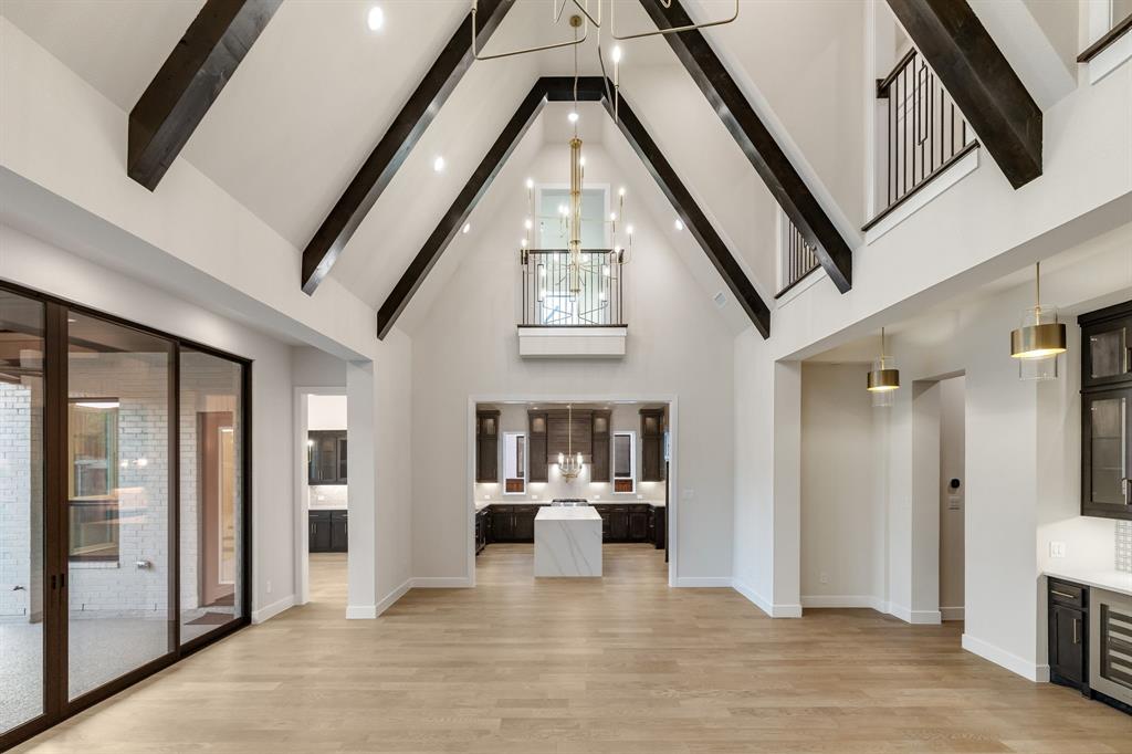 Vaulted ceilings with exposed beams, wood-finish flooring, an interior balcony with metal railing, floor-to-ceiling glass panel doors, and a kitchen island with a stone-finish countertop