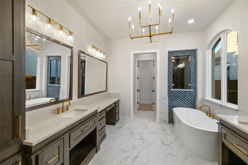 Dual-vanity cabinetry with stone-surface countertops, freestanding soaking tub, arched window, decorative tile accent wall, and brass-finish fixtures
