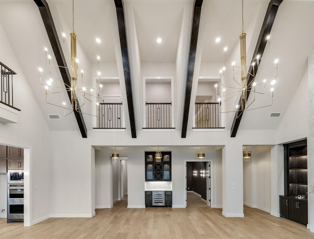 Vaulted ceilings featuring exposed dark-toned beams, dual multi-arm brass chandeliers, wood-finish flooring, built-in cabinetry with integrated wine refrigeration, and interior balcony railings