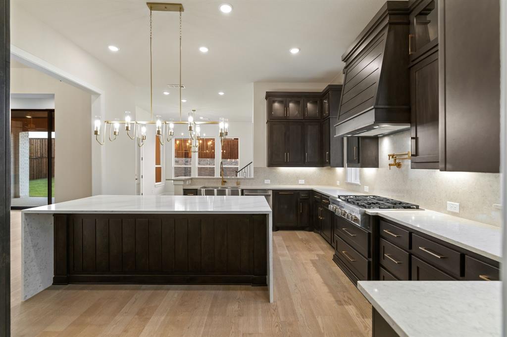 Kitchen featuring a waterfall-edge island, dark-toned cabinetry, a custom range hood, stone countertops, and wood-finish flooring