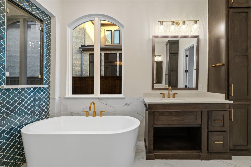 Primary Bath Freestanding soaking tub with a wall-mounted arched window, brass finish fixtures, decorative blue tile backsplash, and dark wood cabinetry with a stone countertop