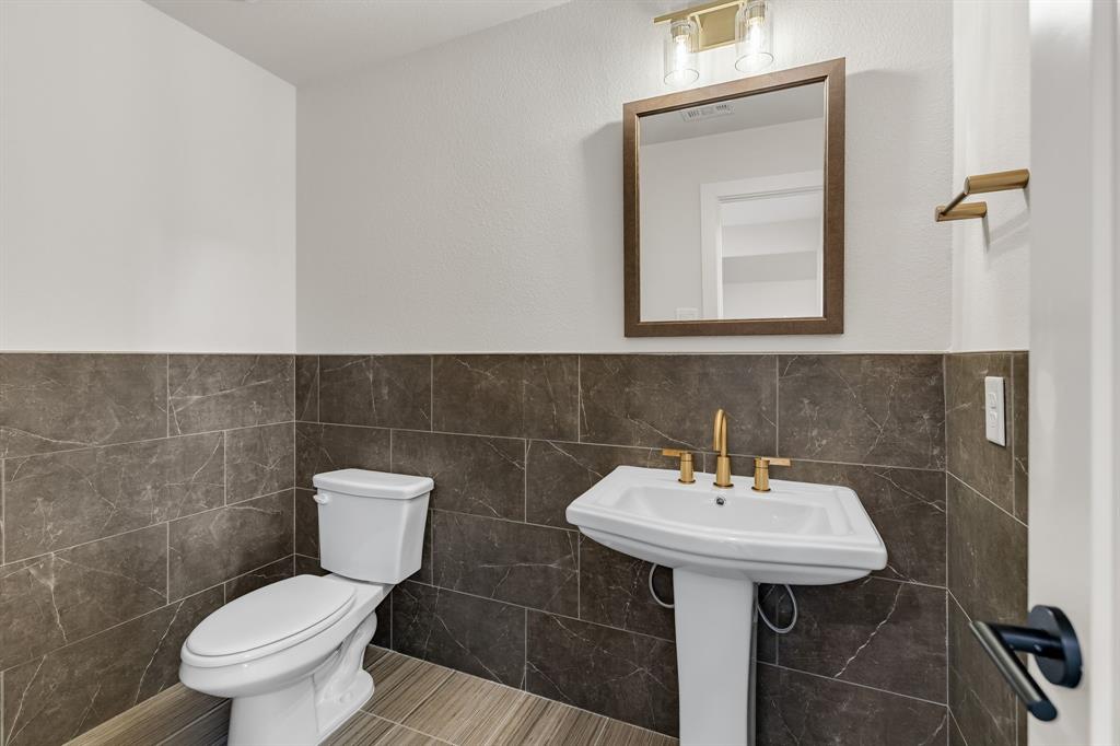 Upstairs Powder room featuring stone-finish wall tile, pedestal sink, gold-tone faucet hardware, framed mirror, and two-light wall sconce