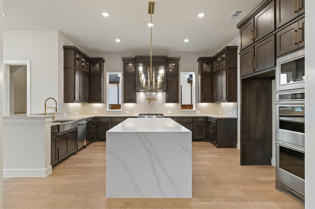 Kitchen featuring dark wood cabinetry, a large center island with a stone-finish waterfall edge, stainless steel appliances including a built-in double oven, wood-finish flooring, and a decorative metallic chandelier