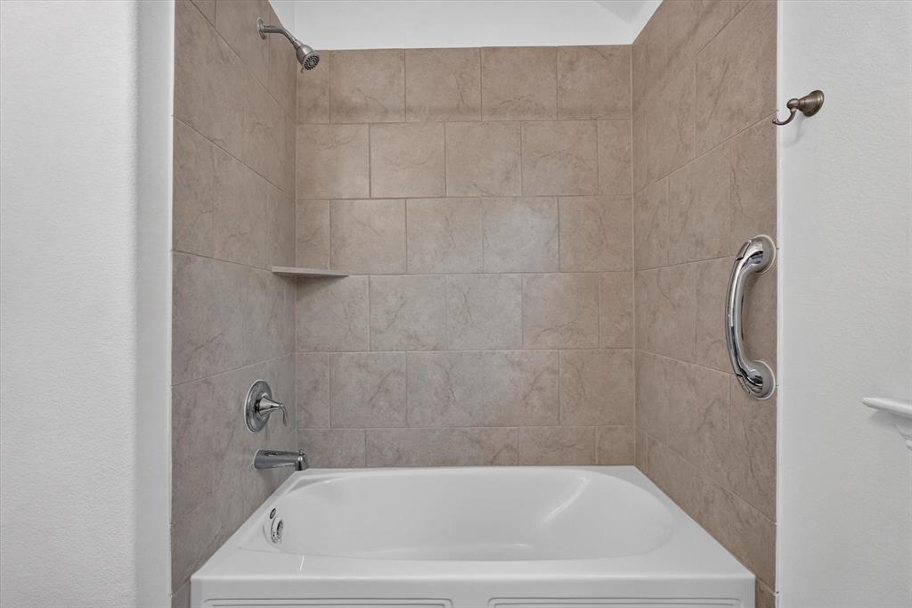 Bathtub alcove featuring neutral-toned tile surround, built-in shelving, chrome fixtures, and a wall-mounted support bar