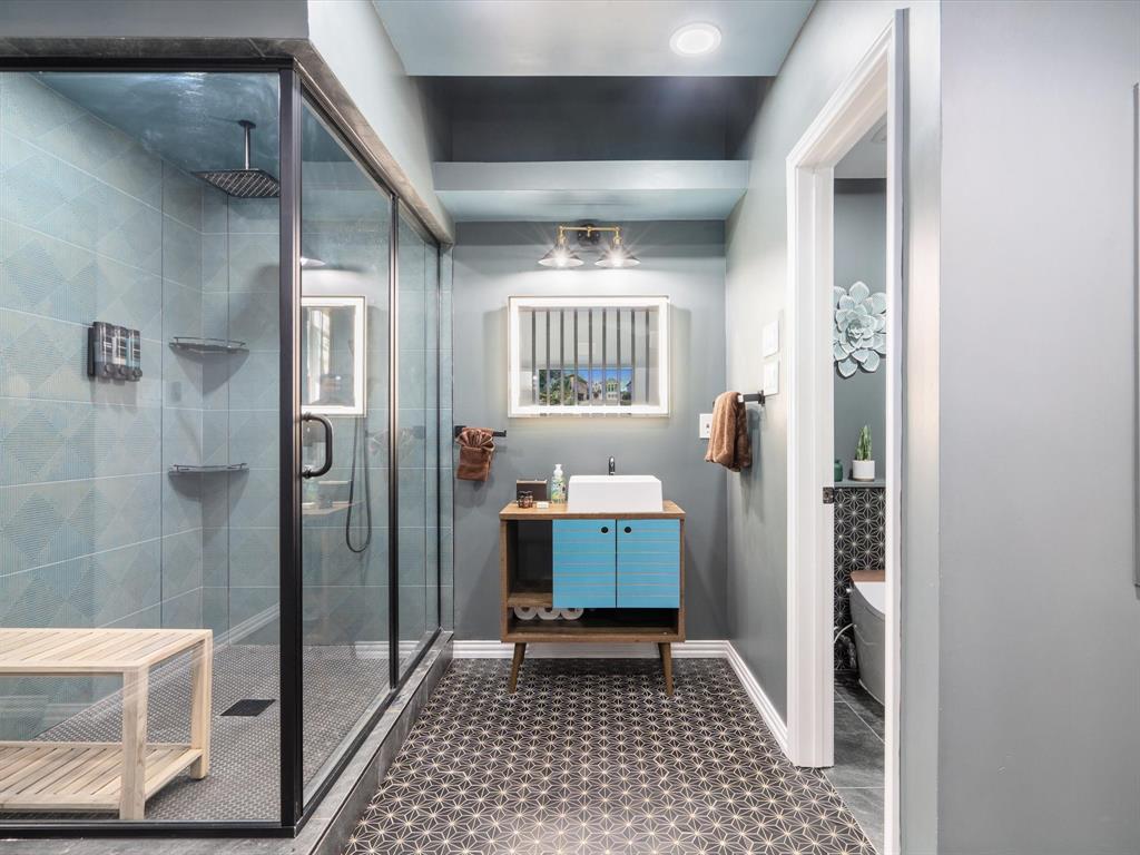 Ensuite Bathroom – In-Law SuiteSecond-floor full ensuite: walk-in glass shower with rain head, gray tile surround/floors, wood vanity with blue cabinetry and vessel sink, recessed lights, wooden bench. Modern & private.