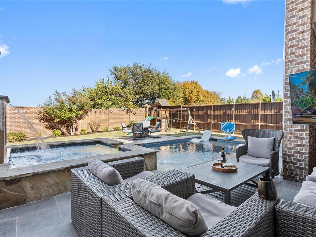 Outdoor living features a stunning backyard oasis with a sparkling swimming pool and attached hot tub, a spacious patio with wicker sectional seating and a coffee table, a covered hangout area, a fenced yard with a playset, mature trees, and a serene fountain. Perfect for relaxation and family fun.